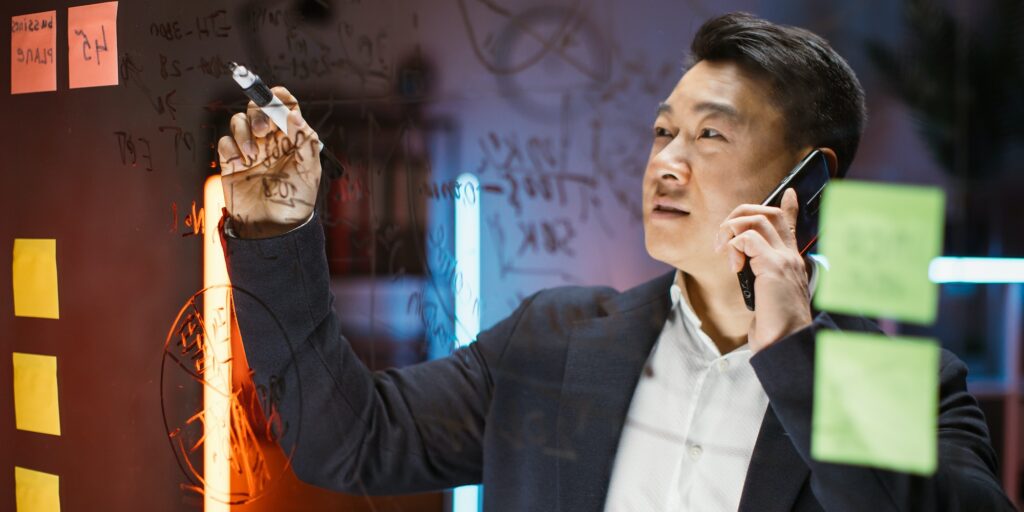 Focused Asian male ceo writing on glass board with sticky notes and talking phone