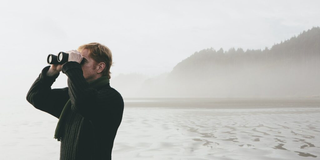 Middle aged man standing on a beach, looking through binoculars at Seabrook, Washington, USA.
