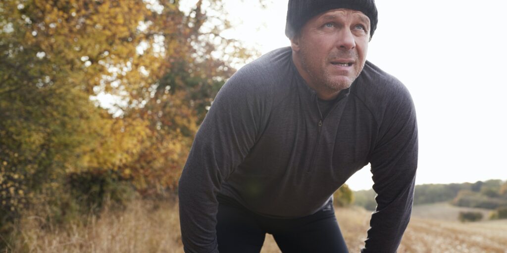 Mature Male Runner Pausing For Breath During Exercise In Woods