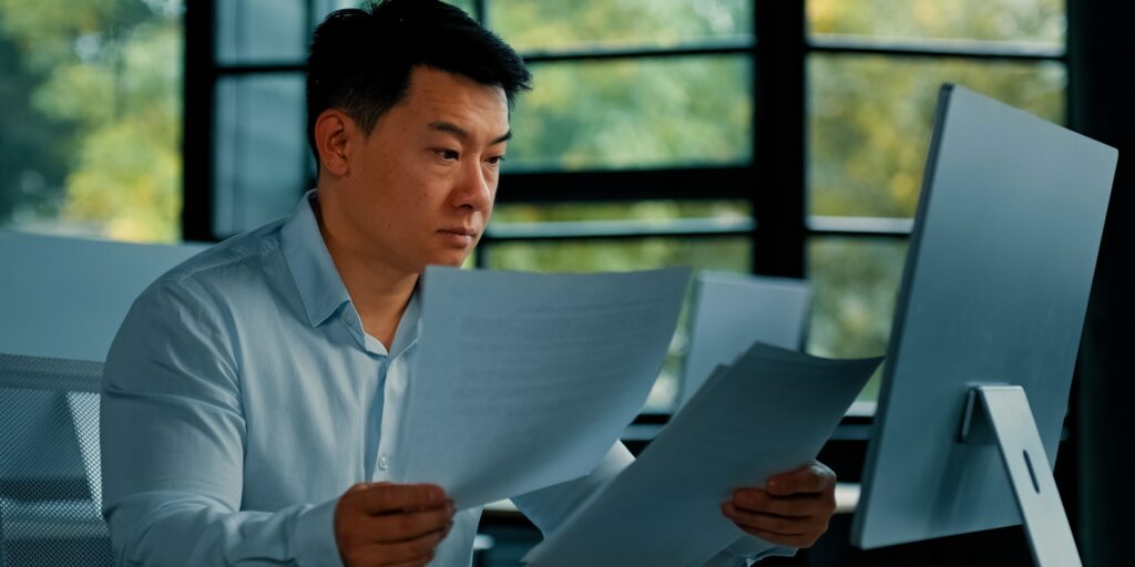 Tired unhappy Asian man worker manager sit at office doing routine paperwork read contract documents
