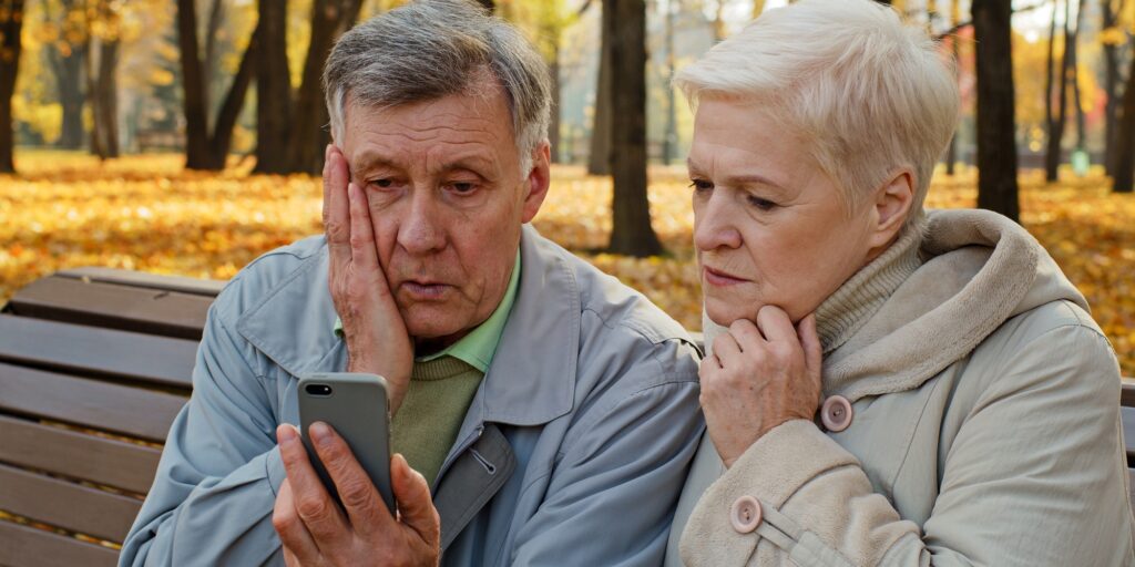 Upset elderly married couple sit on bench in autumn park read bad news on smartphone worried old