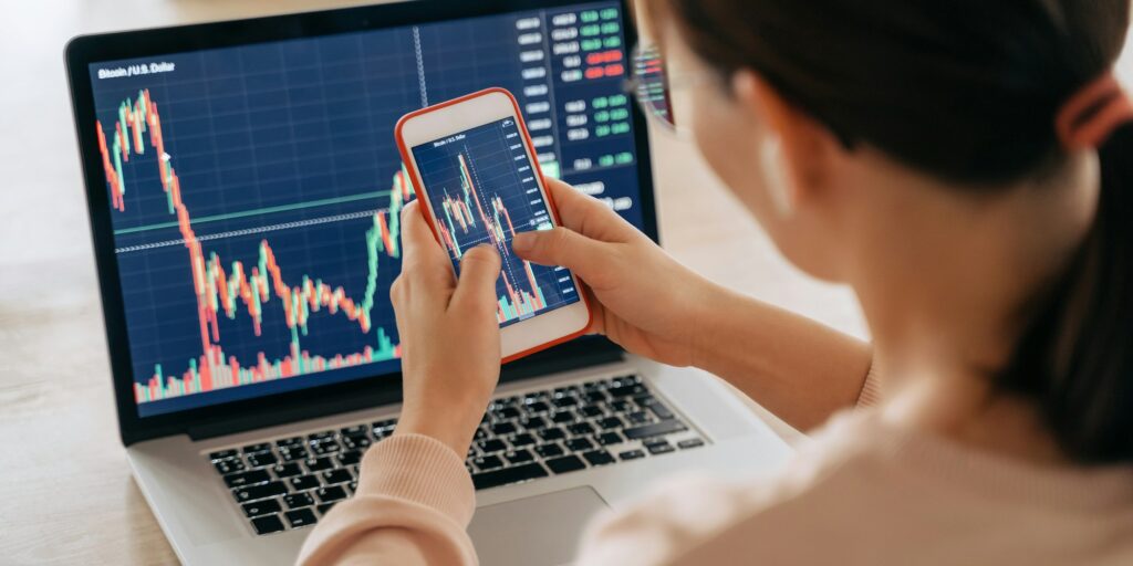 Young female trader investor using smartphone and laptop stock market app for working at home