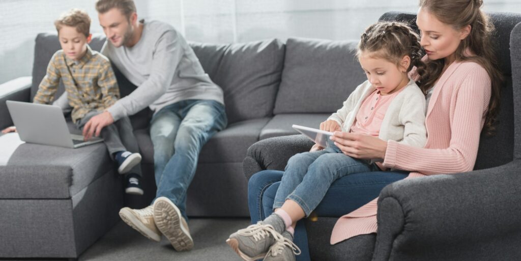 Young family sitting with kids on couch in living room and using digital devices