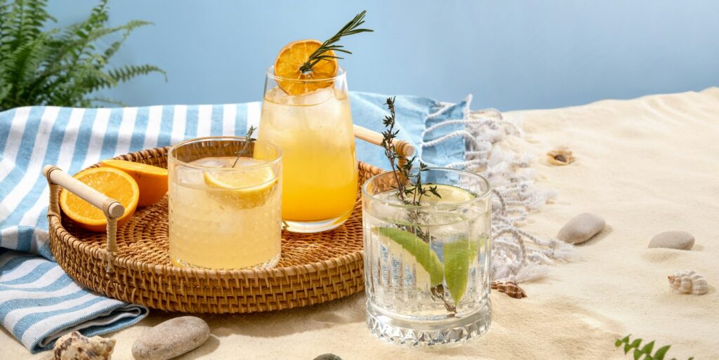 Selection of summer alcoholic cocktails on a wicker tray on beach with white sand.