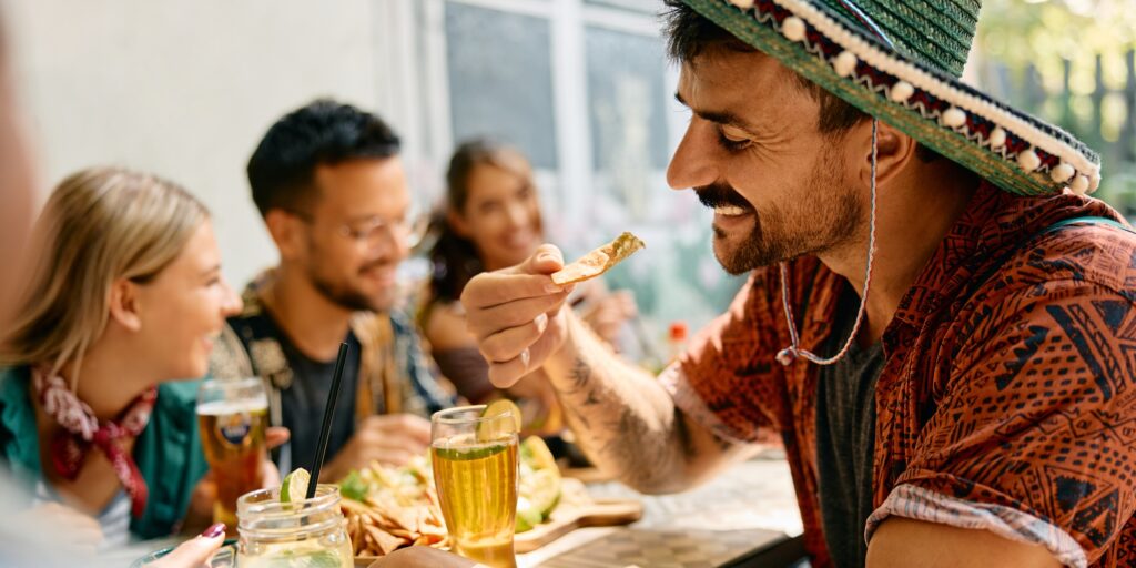 Happy man eating nacho chips while drinking beer with friends in Mexican restaurant.