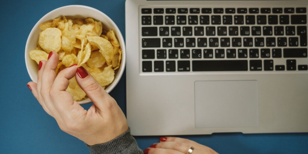 Woman hands with chips and laptop on blue background. Top view