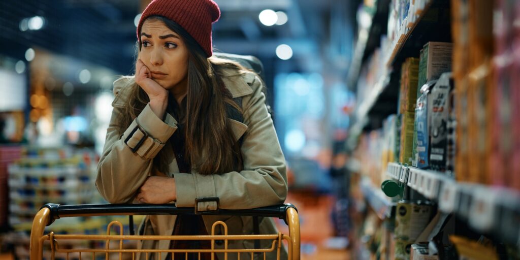 Young sad woman with empty shopping cart among produce aisle at supermarket.