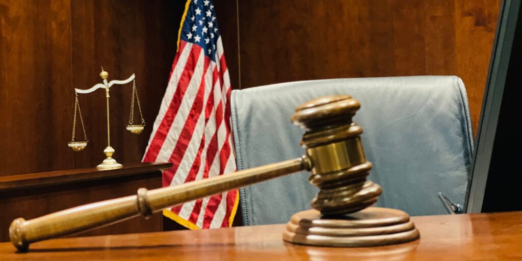 Judge’s wooden gavel on judicial bench, scales of justice and flag of US in background.