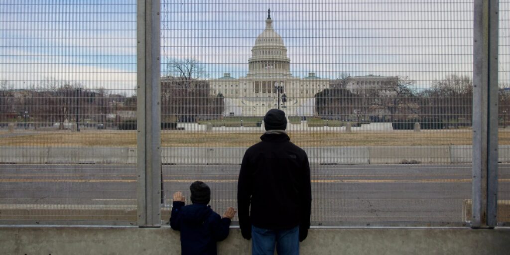 Father and son looking through fence at capitol building after insurrection by Trump supporters