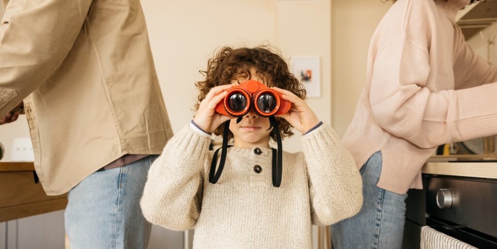 Child playing in a kitchen with toy binoculars amidst his parents.