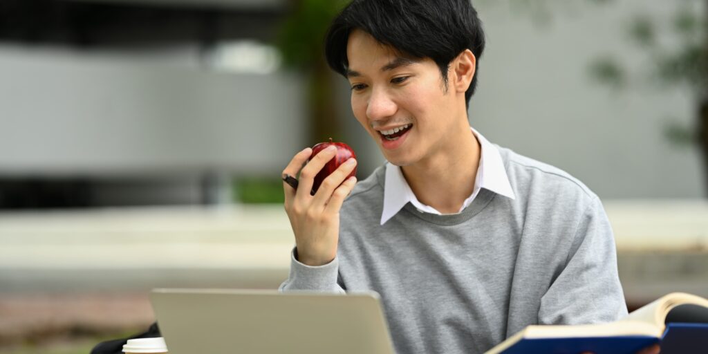 Happy young asian man eating apple and using laptop at campus.